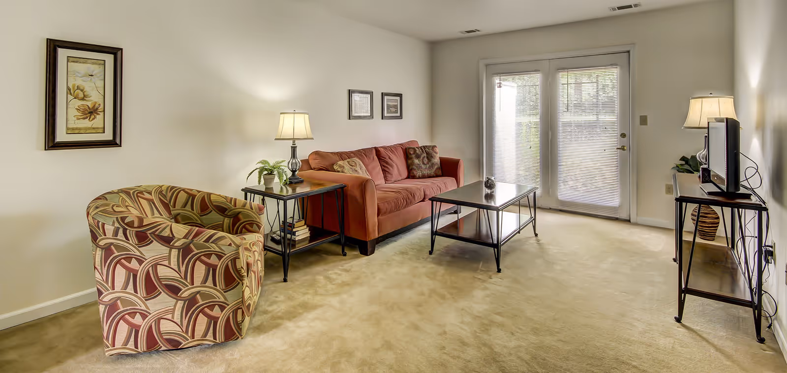 A cozy living room with a patterned armchair, a red sofa with decorative pillows, a glass-top coffee table, and two side tables with lamps. There is a TV on a stand to the right and framed artwork on the walls. A set of glass doors with blinds leads outside.