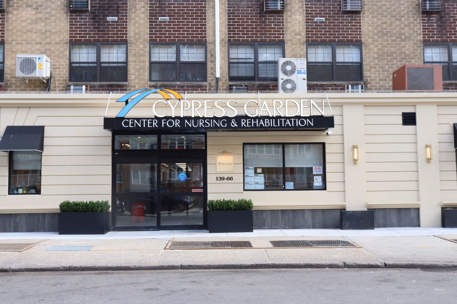 Exterior front view of Cypress Garden Center for Nursing and Rehabilitation building entrance with glass doors, windows, and signage above the entrance. The building has a beige facade with brick upper walls and air conditioning units mounted above.