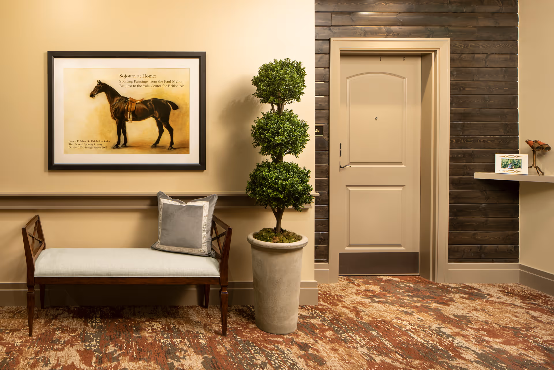 A hallway area in a senior living facility with a closed beige door, a wooden bench with a gray cushion and pillow, a tall potted plant with three round green sections, a framed picture of a horse on the wall, and a small shelf with decorative items on the right side.