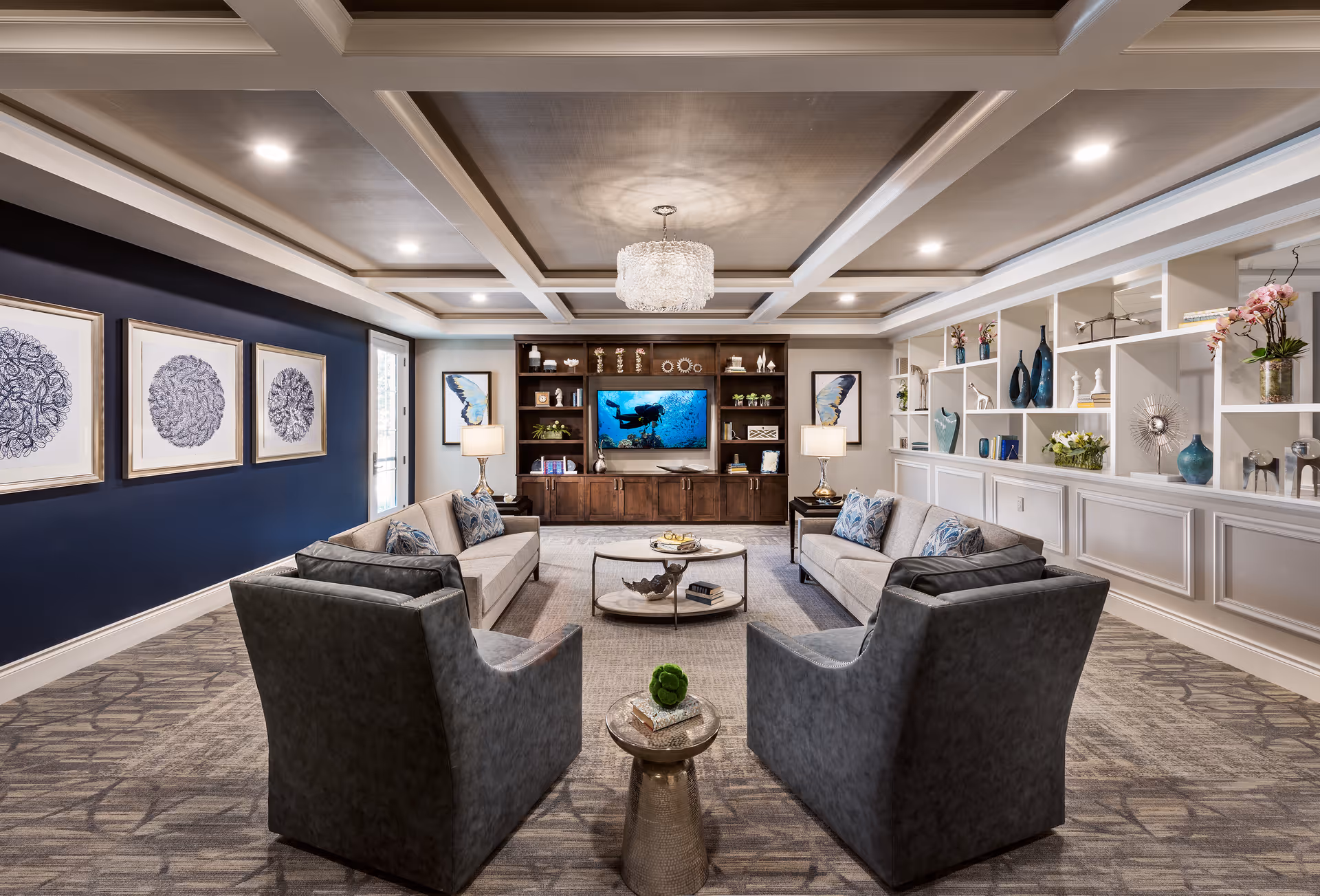Well-appointed communal living room with sofas and armchairs arranged around a coffee table facing a built-in entertainment center and decorative shelving.