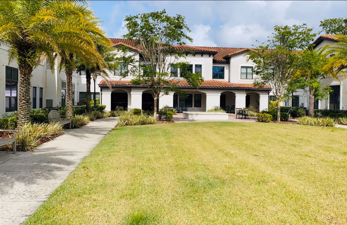 Well-maintained grassy courtyard with a walkway, palm trees and benches in front of a two-story Mediterranean-style building.