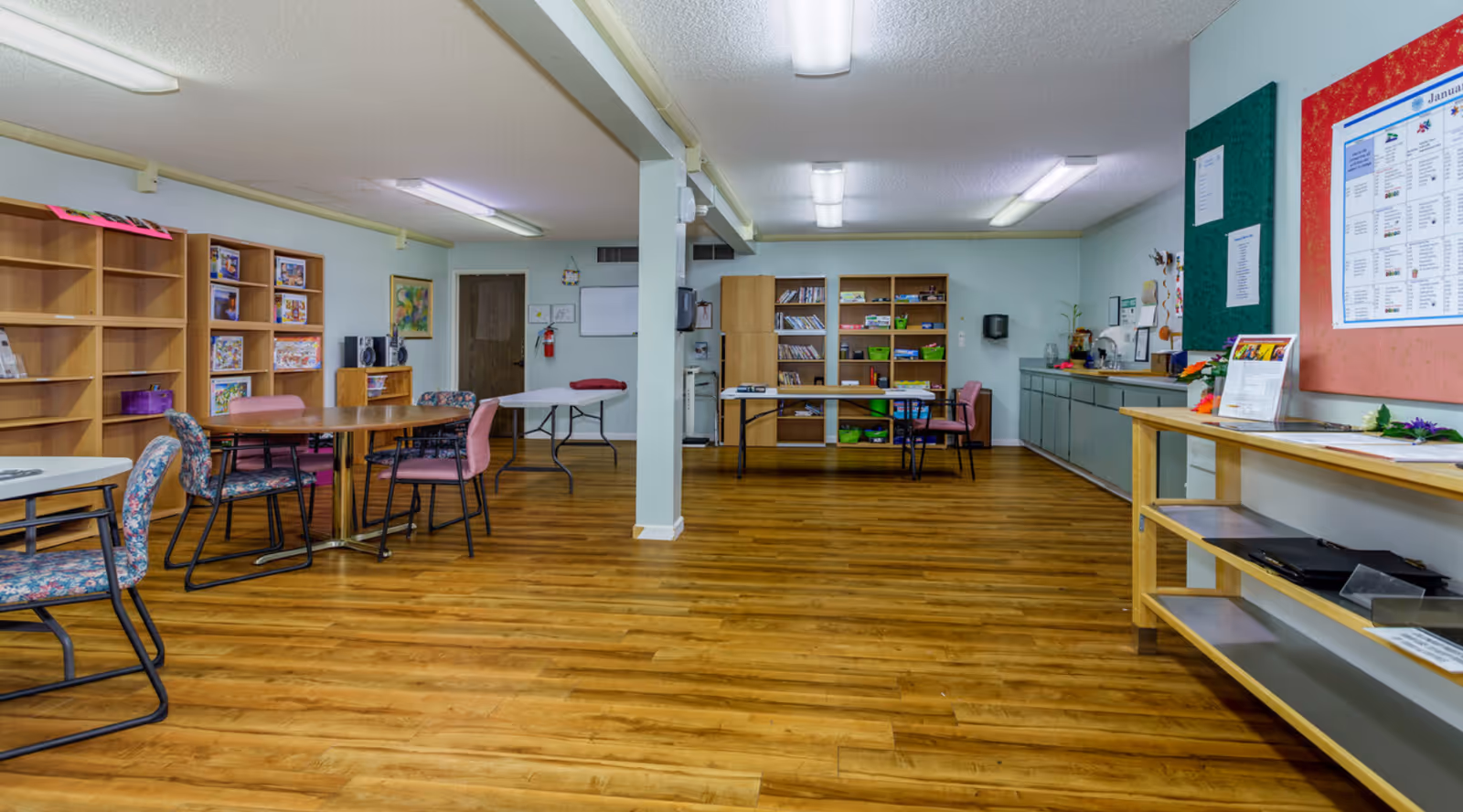 A spacious community room with wooden flooring, multiple tables and chairs, bookshelves filled with games and books, bulletin boards on the walls, and a counter area with cabinets and a sink.