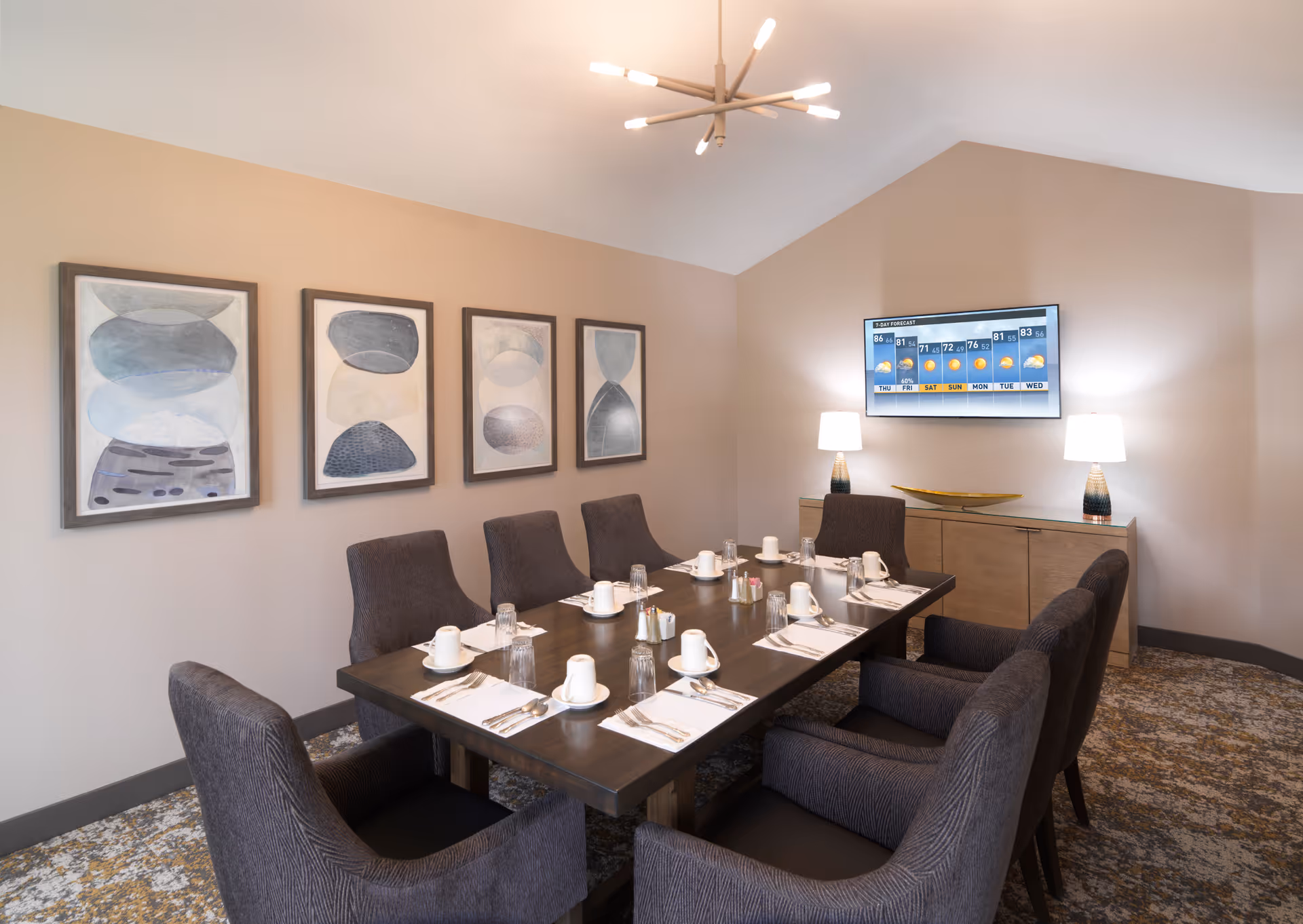 A private dining room with a long set table, upholstered chairs, framed abstract wall art, a sideboard with lamps, and a wall-mounted TV.