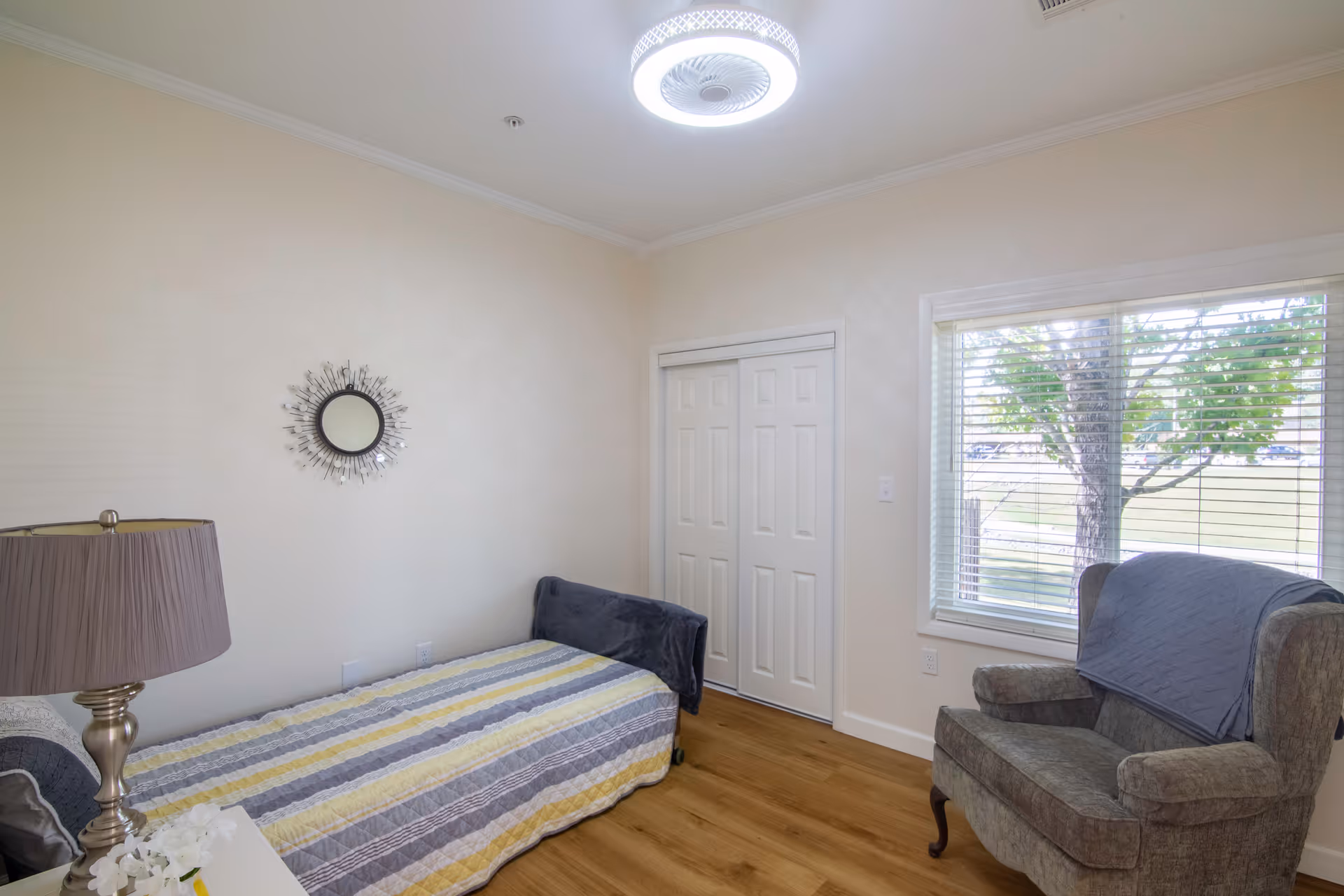 A small bedroom with a single bed covered with a striped yellow, gray, and white blanket. There is a bedside table with a lamp and flowers, a decorative round mirror on the wall, a gray armchair with a blue cover, a window with blinds showing a tree outside, and a closet with white double doors. The room has light-colored walls and wooden flooring.