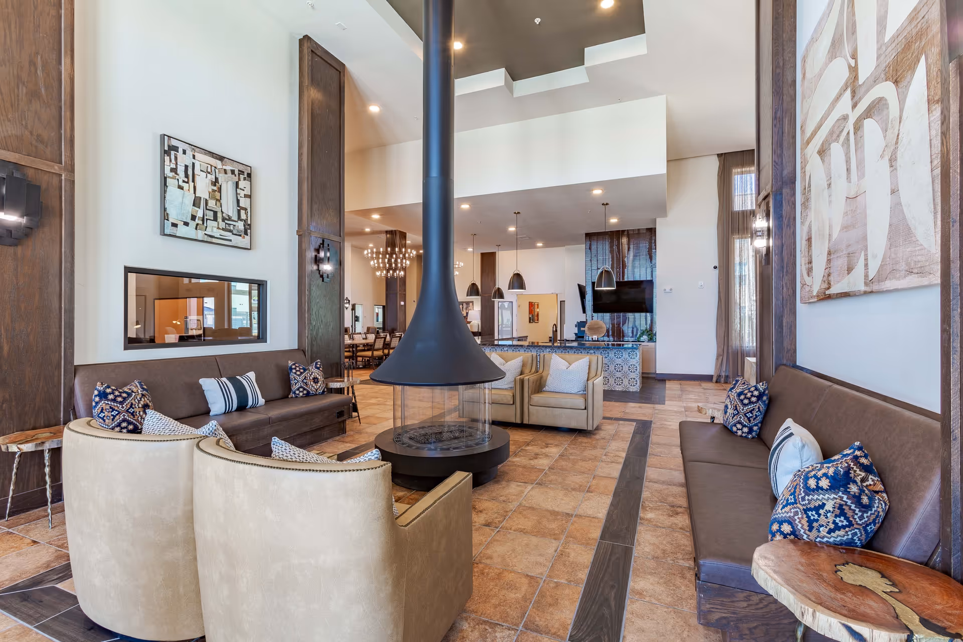 A spacious and modern living room area with a central black suspended fireplace surrounded by beige and brown seating including sofas and armchairs with decorative pillows. The room features tiled flooring with wood accents, large abstract wall art, and pendant lights hanging over a counter in the background. There is a dining area visible further back with a chandelier and multiple chairs around tables.