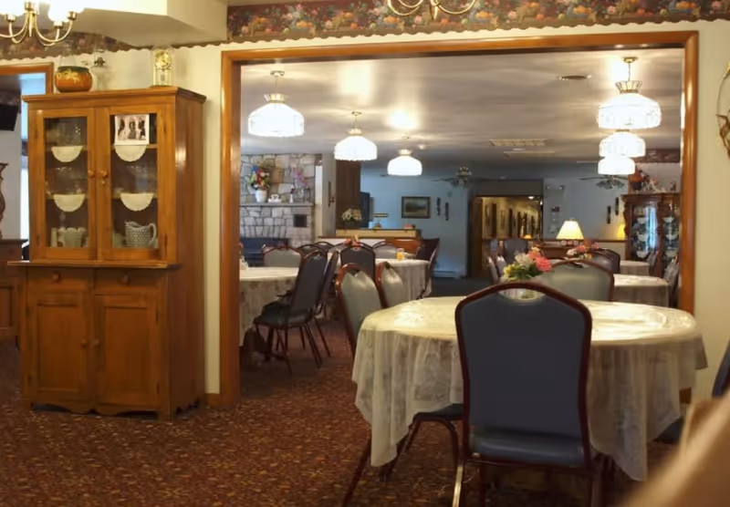 Interior view of a dining room in a senior living facility with round tables covered in lace tablecloths and surrounded by chairs. The room has warm lighting with chandeliers hanging from the ceiling, a wooden cabinet with glass doors displaying dishes, and floral decorations on the tables. A stone fireplace is visible in the background.