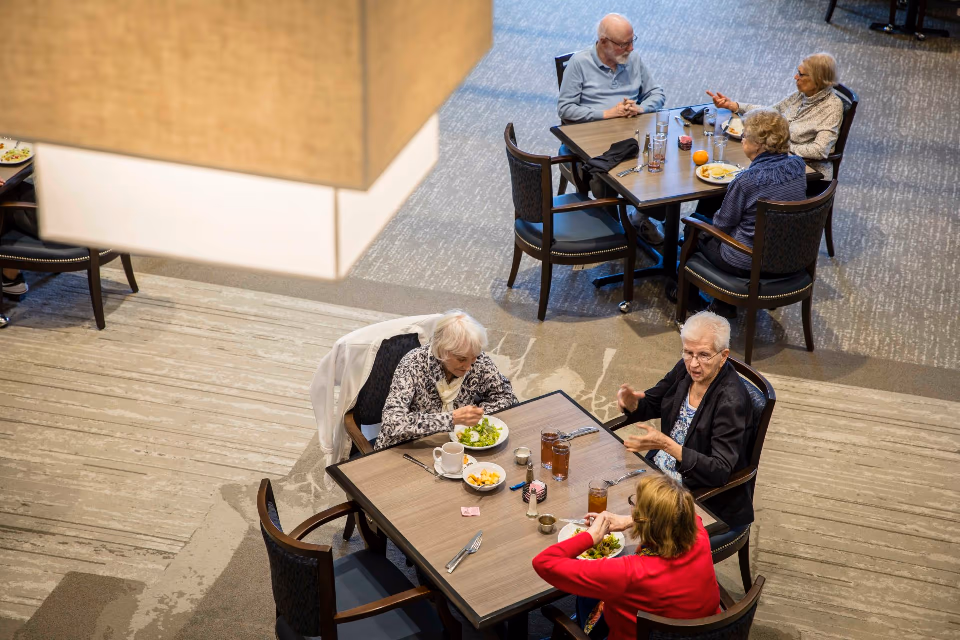 Overhead view of several elderly residents seated at dining tables in a senior living facility, eating and conversing.