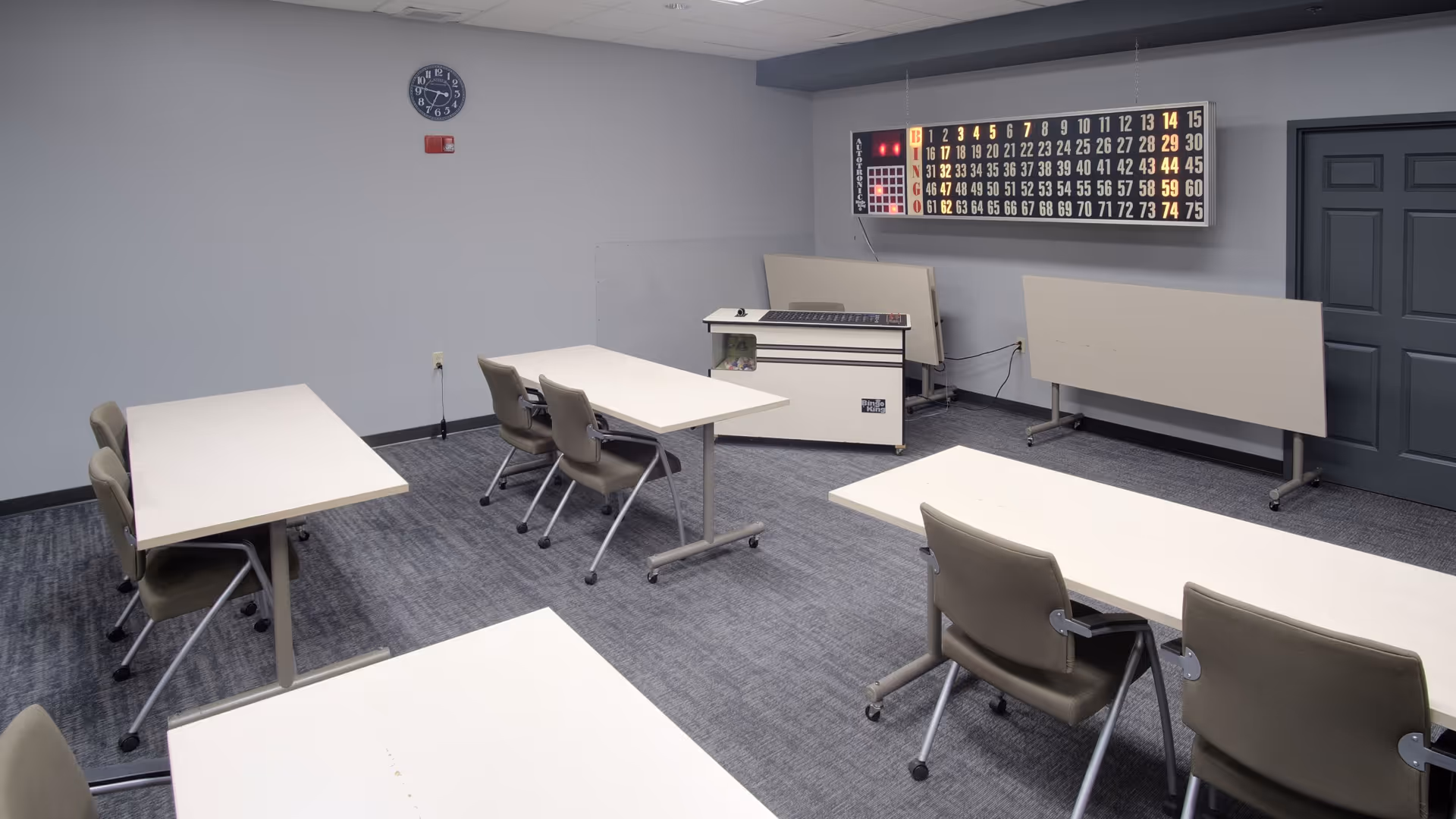 A room with several tables and chairs arranged in rows, a large electronic bingo board on the wall, a clock, and a podium or desk at the front. The room has gray walls and carpeted floor, with a closed door on the right side.