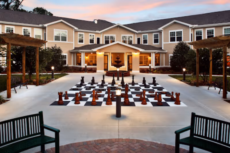 Outdoor courtyard area of a senior living facility at sunset featuring a large outdoor chessboard with oversized chess pieces, benches, pergolas, and a central fountain in front of a two-story beige building with multiple windows.