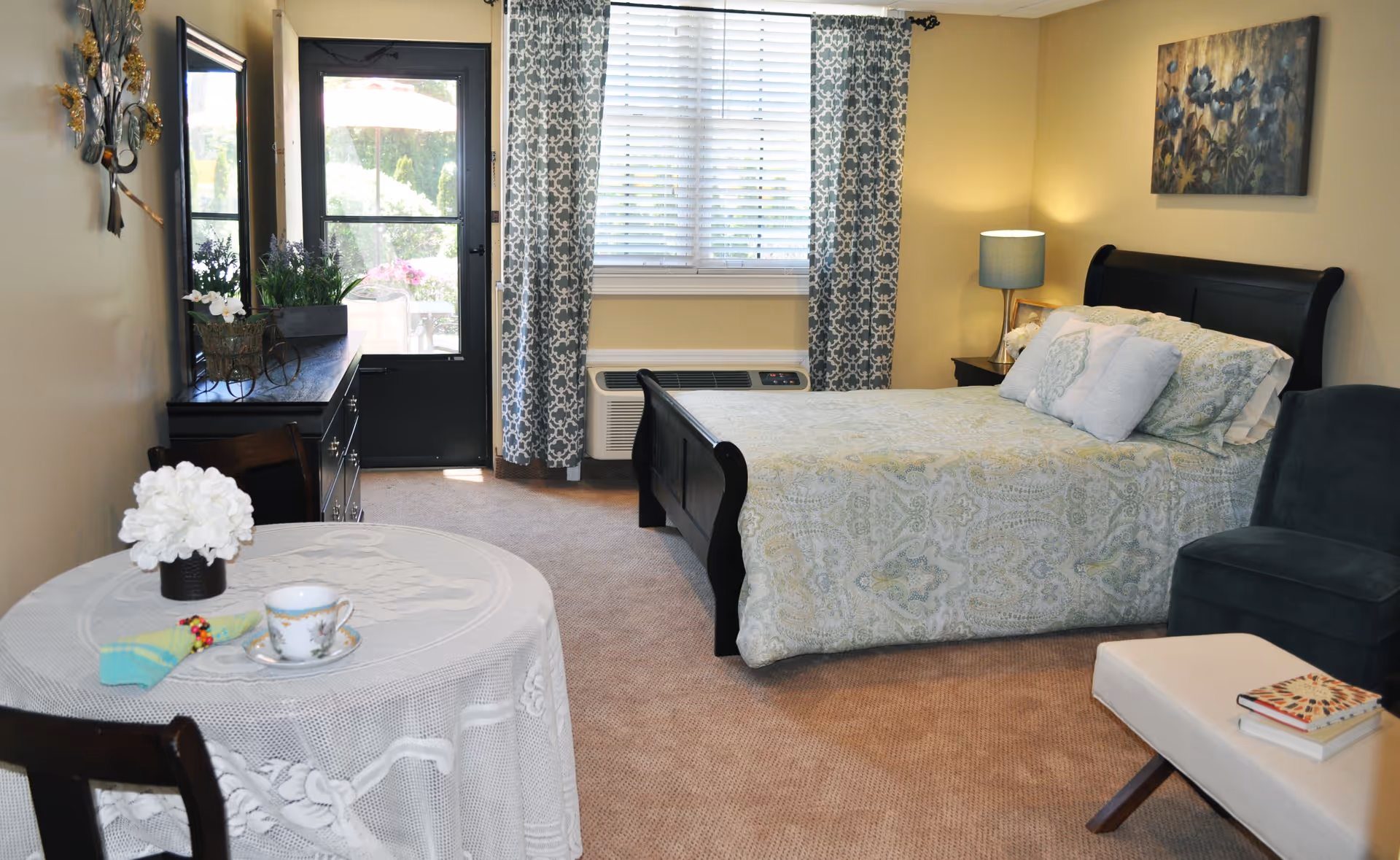 A cozy bedroom in an assisted living facility featuring a bed with patterned bedding, a nightstand with a lamp, a small round table with a lace tablecloth and a teacup, a dresser with a mirror, a window with patterned curtains, and a door leading outside. There is also a comfortable chair and an ottoman with books on it.