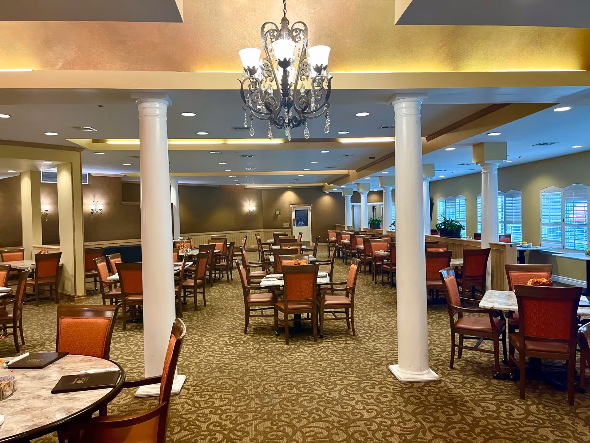 Spacious dining room with multiple tables and chairs arranged neatly. The room features white columns, a decorative chandelier, wall sconces, and windows with white shutters. The carpet has a patterned design, and menus are placed on some tables.