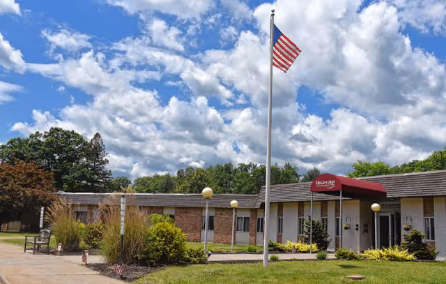 Exterior view of Highview of Northampton facility with a single-story building, an American flag on a flagpole, a red awning over the entrance, and landscaped greenery under a partly cloudy sky.