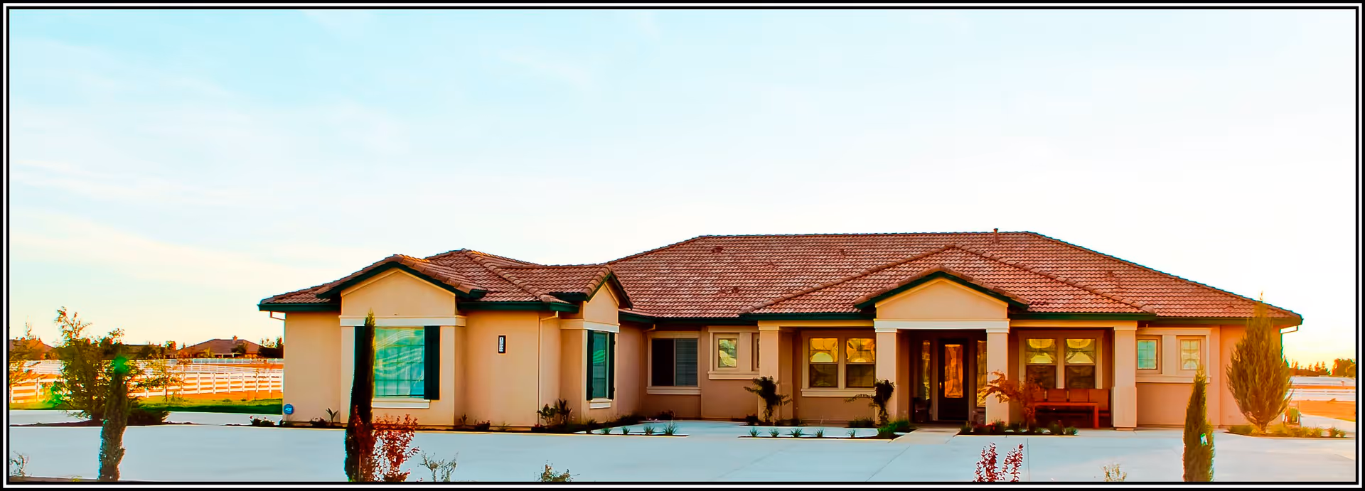 Single-story building with a red-tiled roof and beige exterior walls, surrounded by a paved driveway and small landscaped plants, under a clear sky.
