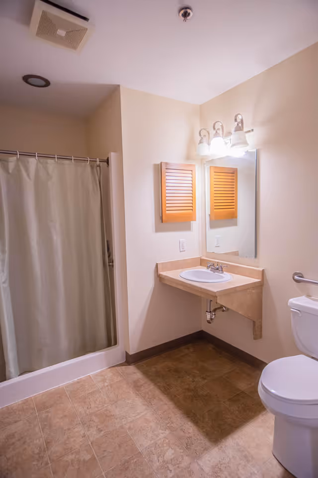 A clean bathroom with a shower area enclosed by a curtain, a wall-mounted sink with a mirror and light fixtures above it, a wooden cabinet with louvered doors, and a toilet. The floor is tiled and the walls are painted a light beige color.