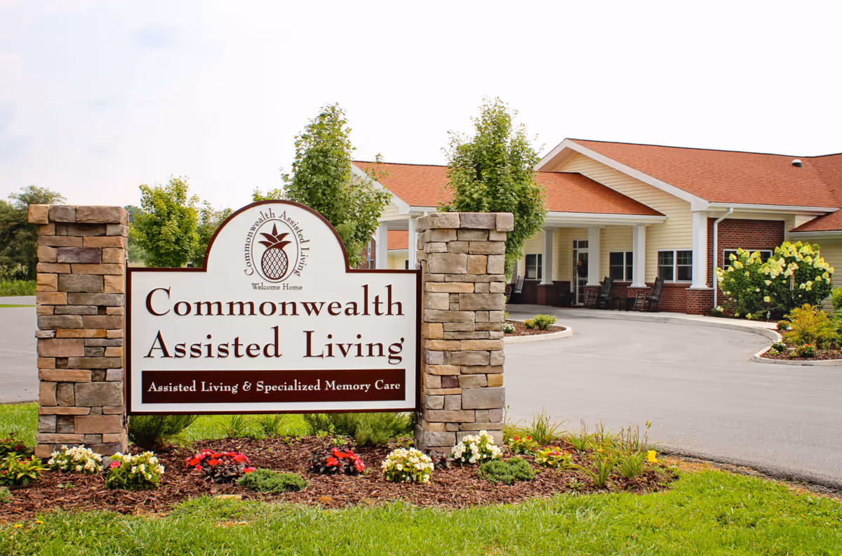 Entrance sign for Commonwealth Assisted Living with a stone pillar on each side, surrounded by flowers and greenery. In the background, there is a single-story building with a red roof, beige siding, and a covered porch with rocking chairs.