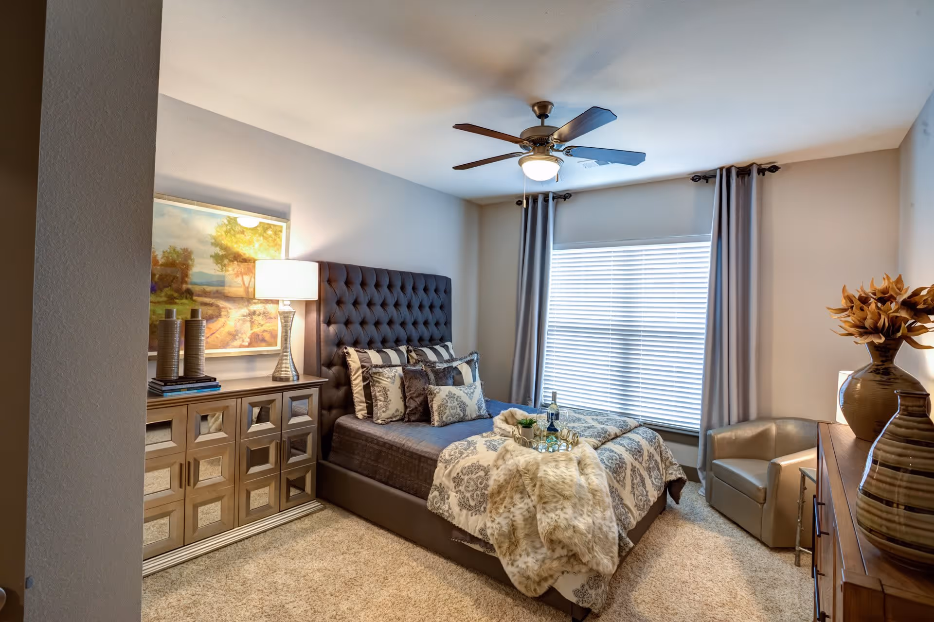 A cozy bedroom with a tufted dark headboard bed adorned with patterned pillows and a decorative throw blanket. There is a wooden dresser with a lamp and decorative items on the left side, a large window with closed blinds and curtains behind the bed, a ceiling fan with a light, and a small armchair next to a wooden chest with vases on the right side.