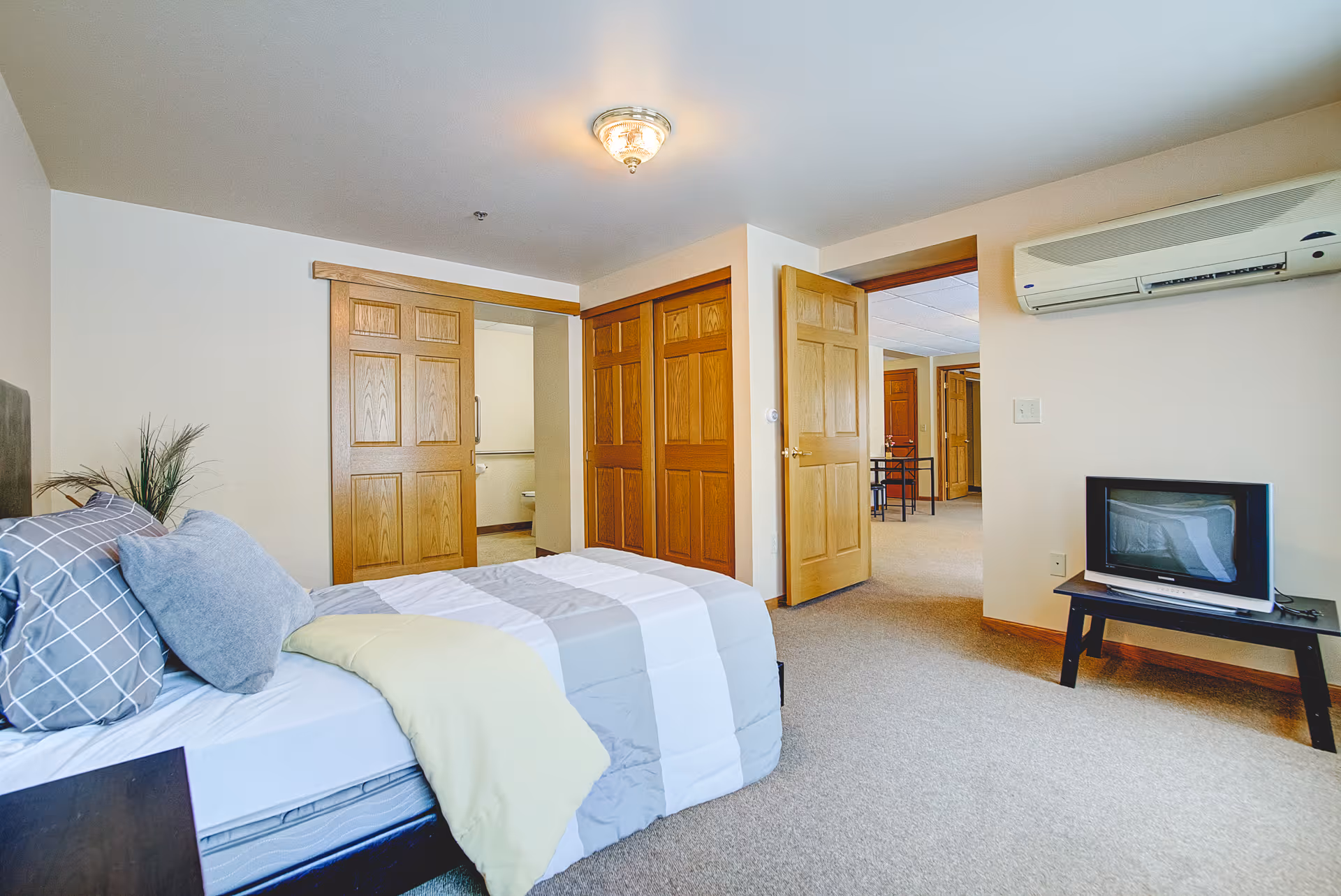 Bright bedroom with a made bed, pillows, wooden doors, a small TV on a stand and a wall-mounted air conditioner.