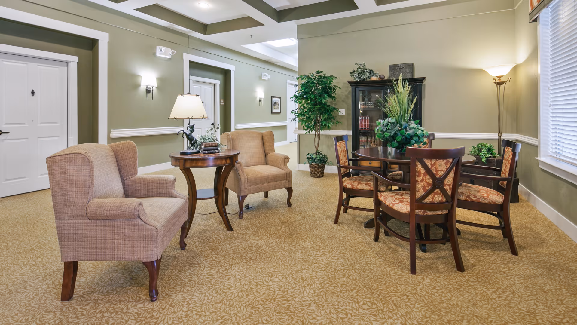 A cozy sitting area in an assisted living facility featuring two beige upholstered armchairs around a small wooden side table with a lamp and decorative items. Nearby is a round wooden table with four patterned cushioned chairs. The room has green walls with white trim, a carpeted floor with a floral pattern, a tall floor lamp, a window with blinds, and a cabinet with plants and books.