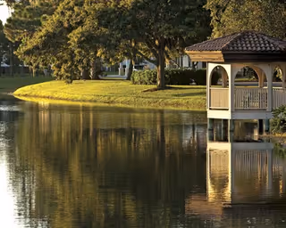 A small wooden gazebo on the edge of a pond reflected in calm water with trees and a grassy shoreline.