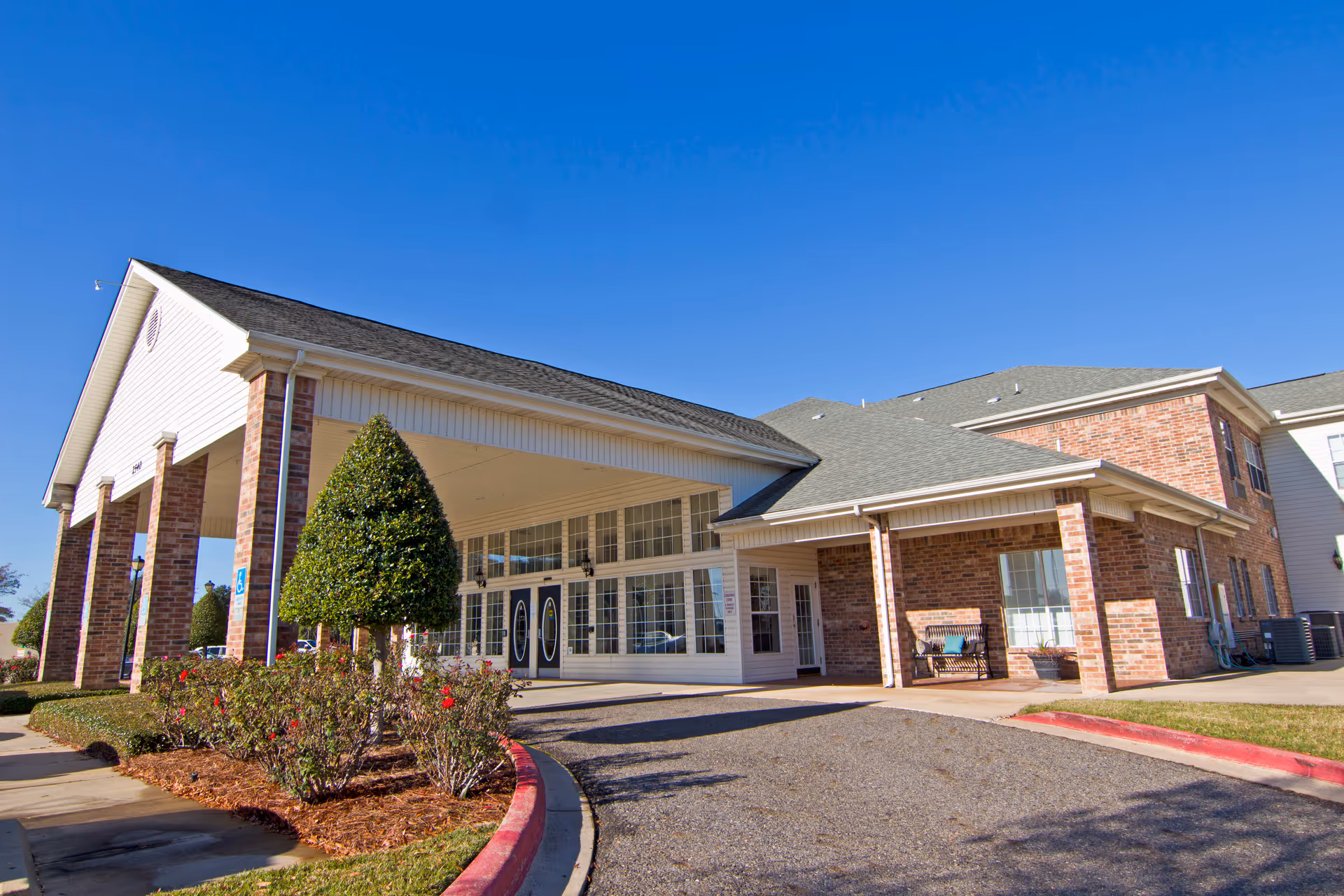 Exterior view of Brookdale Bossier City facility showing a brick building with a covered entrance, manicured bushes, and a clear blue sky.
