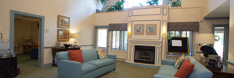 Bright communal living room with two teal sofas facing a central fireplace beneath tall windows and decorative accents.