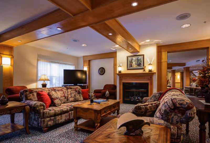 A cozy living room in a senior living facility featuring patterned sofas with red cushions, a wooden coffee table, a fireplace with a framed painting above it, a flat-screen TV, and warm lighting from wall sconces and a table lamp. The room has wooden ceiling beams and carpeted flooring.