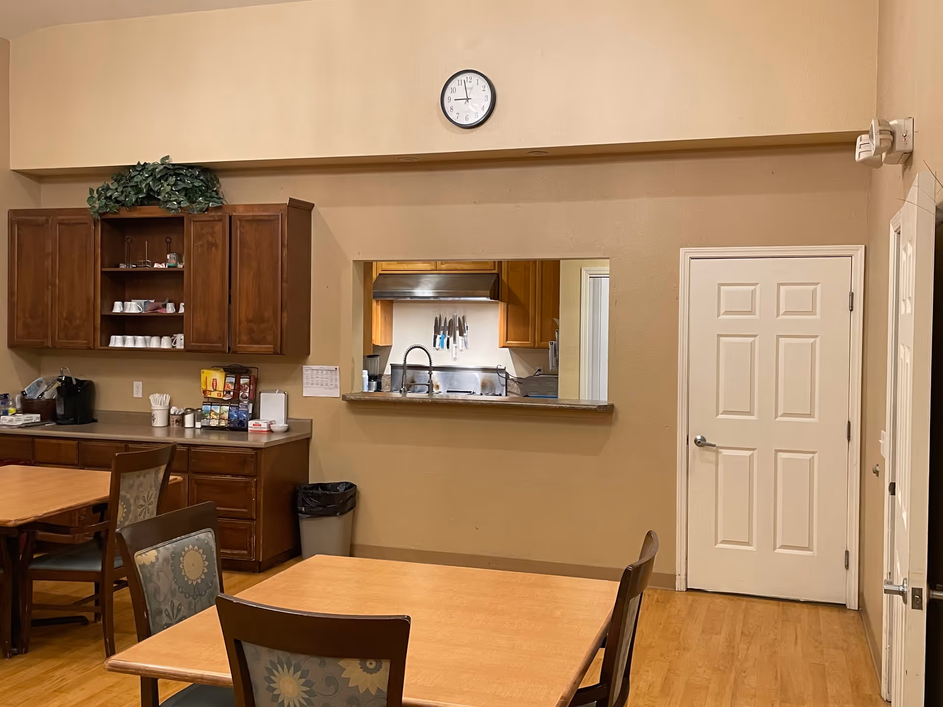 Interior view of a senior care facility dining area with wooden tables and chairs. There is a counter with cabinets above it on the left side, a coffee machine, and various supplies. A pass-through window reveals a kitchen area with knives hanging on the wall. A clock is mounted on the beige wall above the pass-through window, and a closed white door is visible on the right side.
