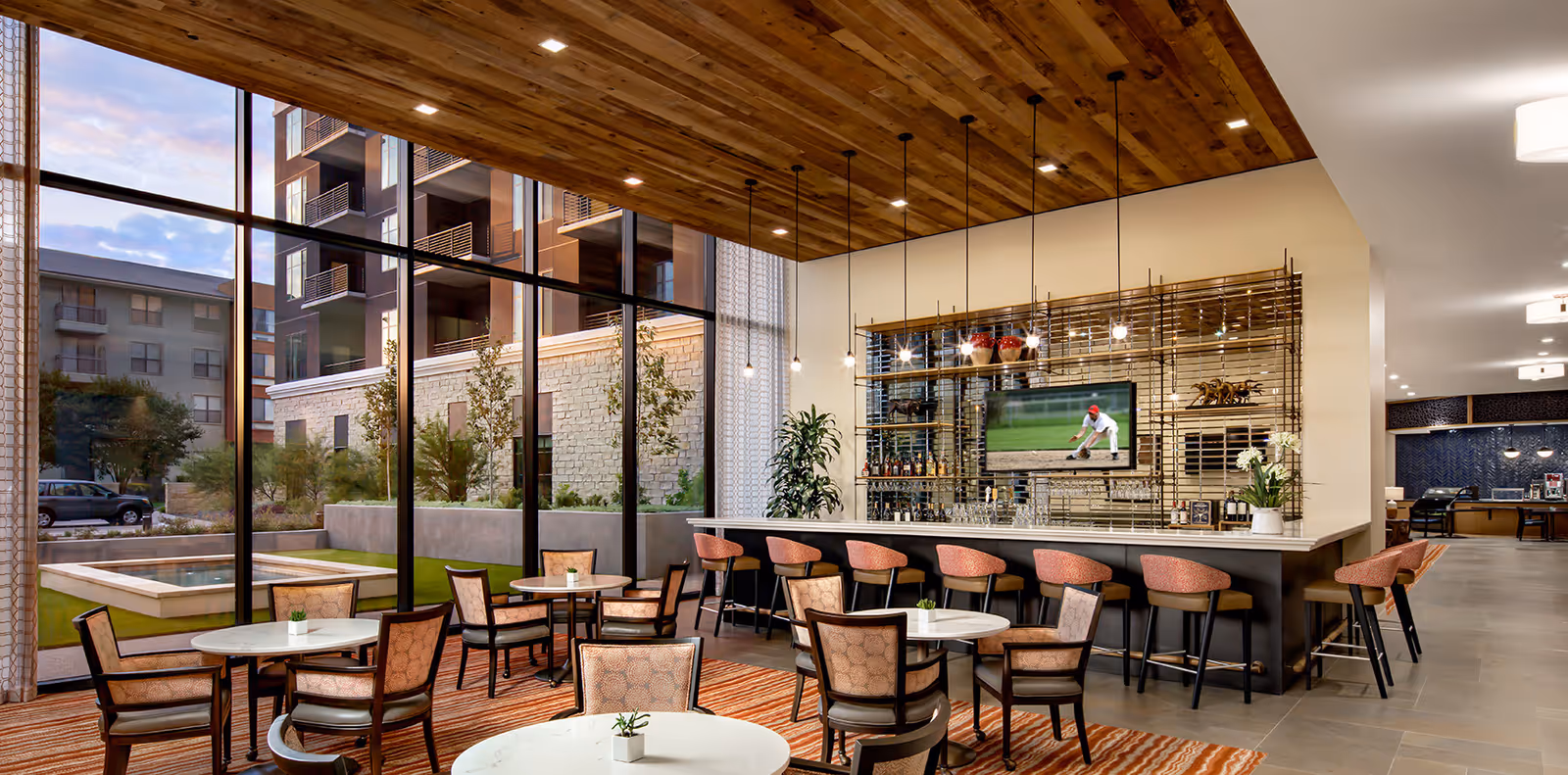 A modern lounge area with a bar and several round tables surrounded by chairs. The bar has high stools and a television mounted on the wall showing a baseball game. Large floor-to-ceiling windows provide a view of the outdoor area and neighboring buildings. The ceiling above the bar is wooden with recessed lighting and hanging pendant lights.