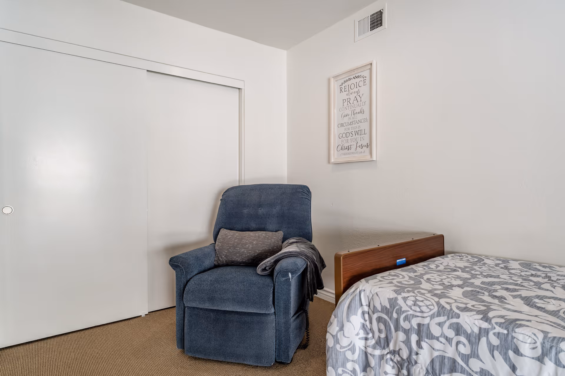 A small bedroom corner with a blue recliner next to a bed with patterned bedding and closed sliding closet doors.