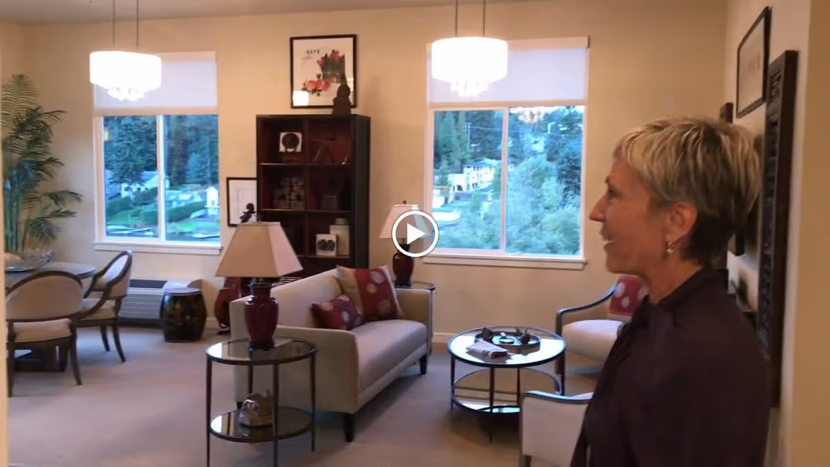 A cozy living room area in a senior living facility with a beige sofa, armchairs, a round coffee table, side tables with lamps, a bookshelf, and large windows showing a green outdoor view. A woman with short blonde hair is standing on the right side of the room.