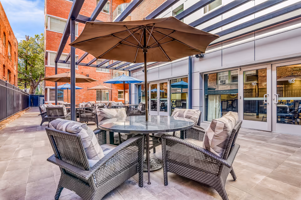 Outdoor patio area at Hollywood Hills Senior Living with multiple round tables surrounded by cushioned wicker chairs and large umbrellas providing shade. The patio is paved with tiles and adjacent to a modern building with large glass doors and windows. There are brick walls and a metal fence enclosing the space.