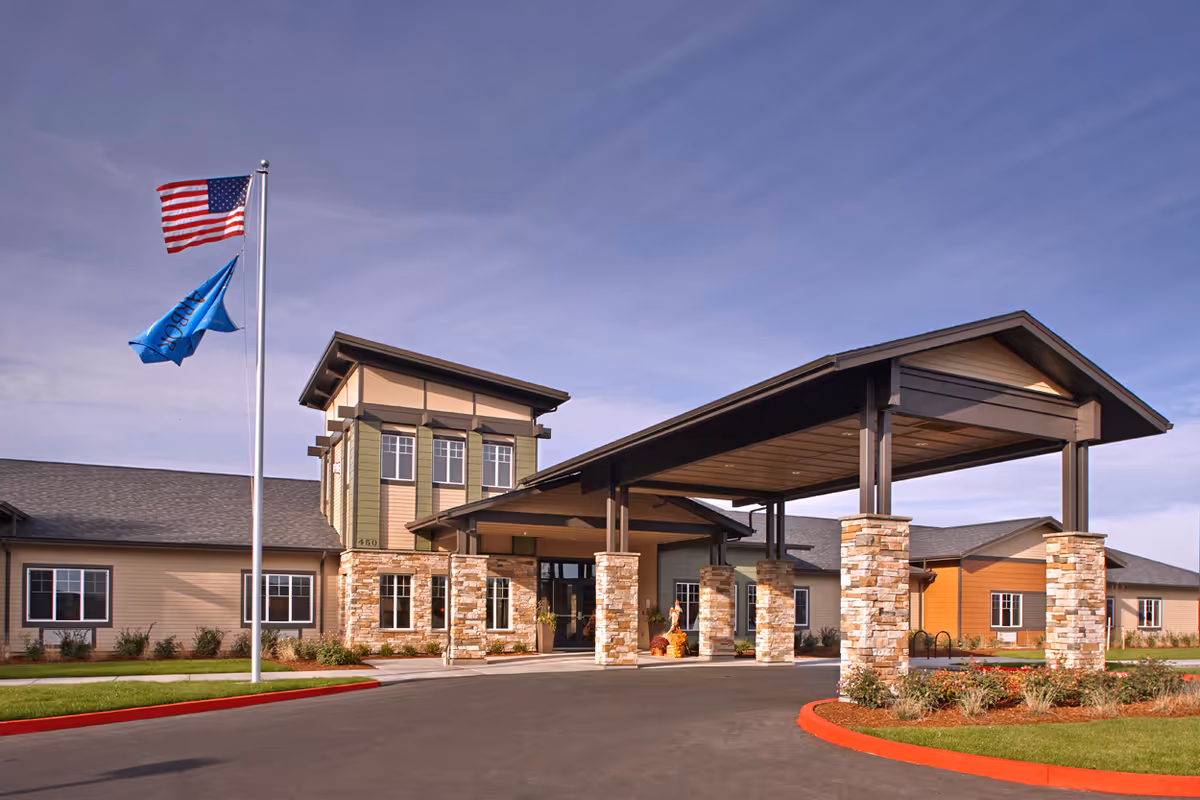 Front entrance of a senior living building with a covered porte-cochere, stone pillars, and two flags flying.