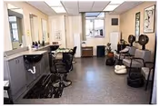 Interior view of a hair salon area in a senior living facility with styling chairs, sinks, and hair dryers. There are chairs lined up against the wall, framed artwork, and a window letting in natural light.