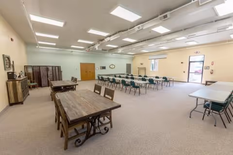 A spacious, well-lit room with multiple tables and chairs arranged for group seating. The room has beige walls, carpeted floor, fluorescent ceiling lights, and a few windows. There is a wooden cabinet and a folding screen along one wall, and an exit door with a window on the far side.