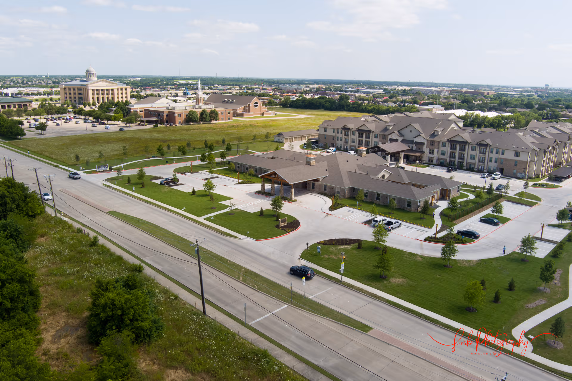 Aerial view of Village Green Memory Care Community Rockwall showing multiple buildings with parking lots, green lawns, and surrounding roads under a partly cloudy sky.