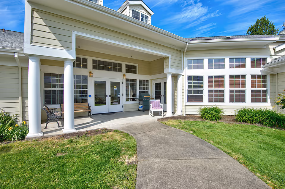 Front entrance of a light-colored assisted living building with white columns, benches, large windows, and a paved walkway through a grassy lawn.