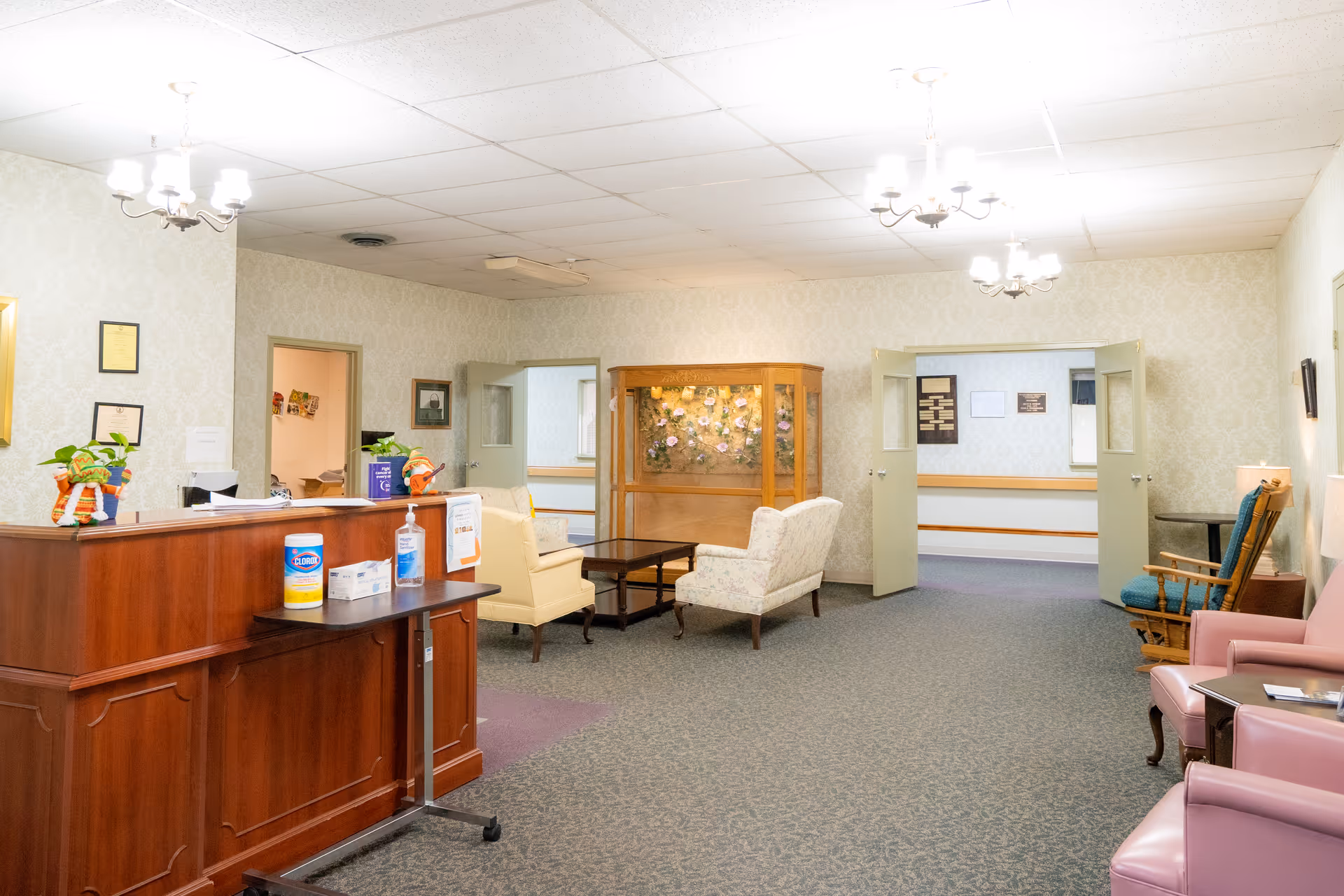 Reception and seating area of a nursing home with a wooden front desk, chairs and couches, a display cabinet, and open double doors.