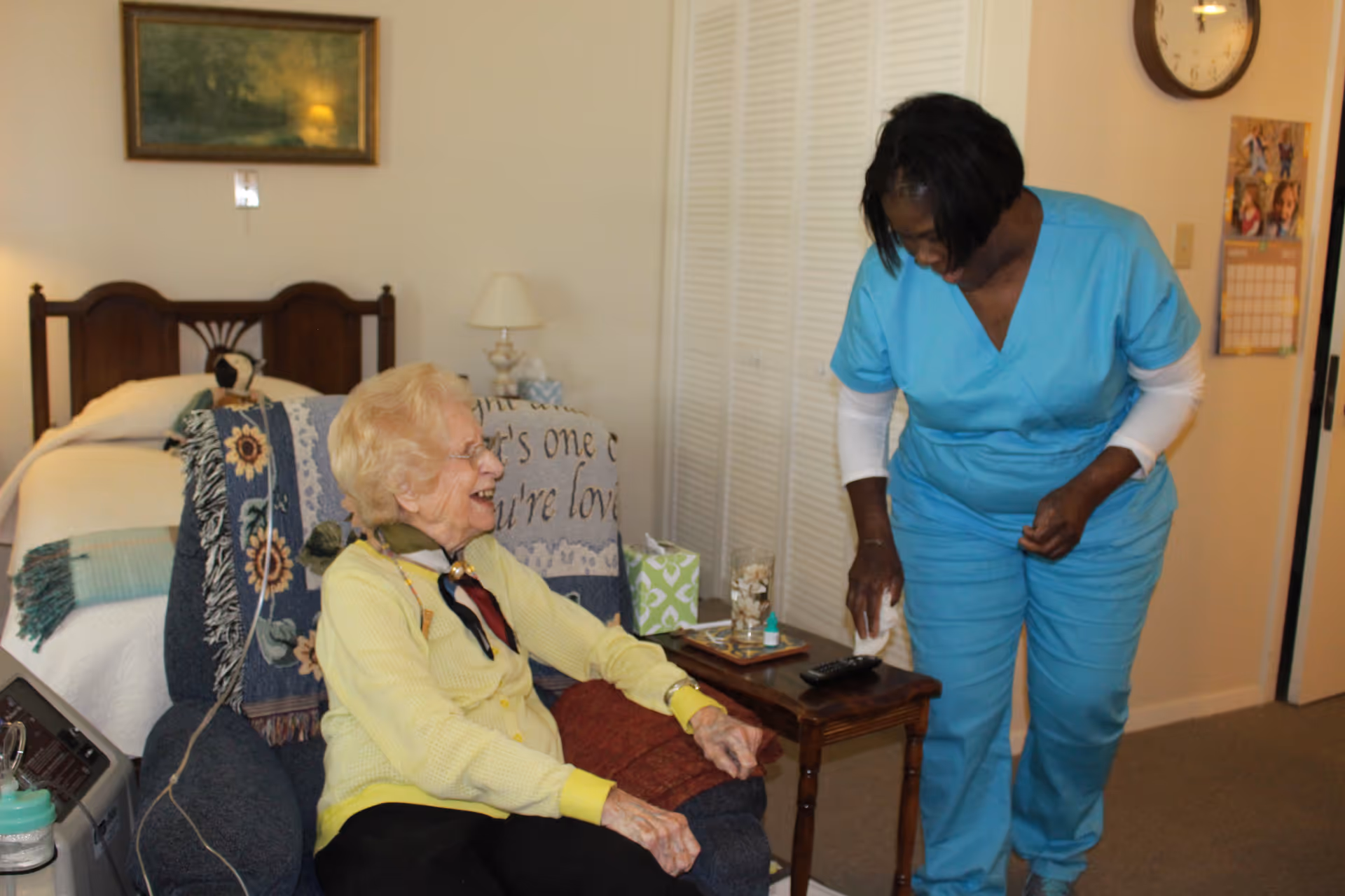 An elderly woman sitting in a cushioned chair covered with a patterned blanket, smiling and interacting with a caregiver dressed in blue scrubs in a cozy bedroom setting with a bed, lamp, and framed picture on the wall.