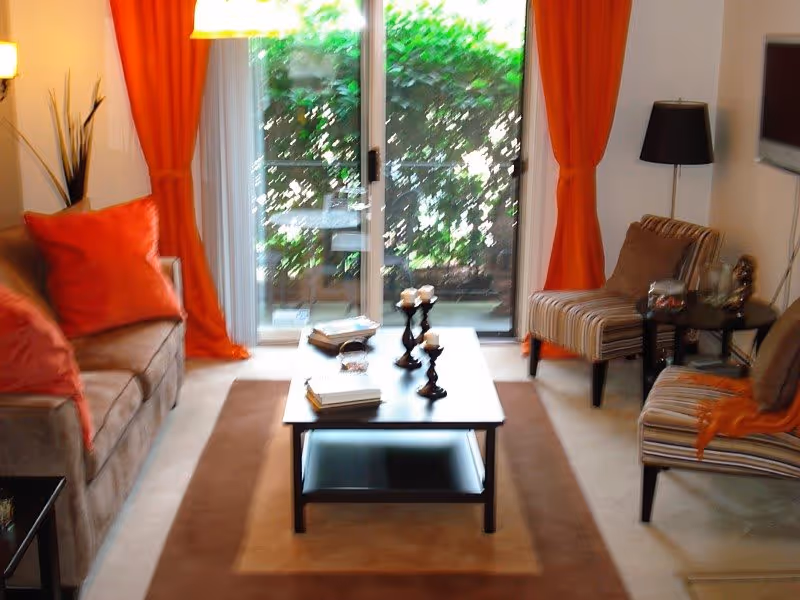 A cozy living room with a brown sofa adorned with orange pillows on the left, two striped armchairs on the right, a dark wooden coffee table with candles and books in the center, orange curtains framing a glass sliding door that opens to a green outdoor area, and a floor lamp and a wall-mounted TV on the right wall.