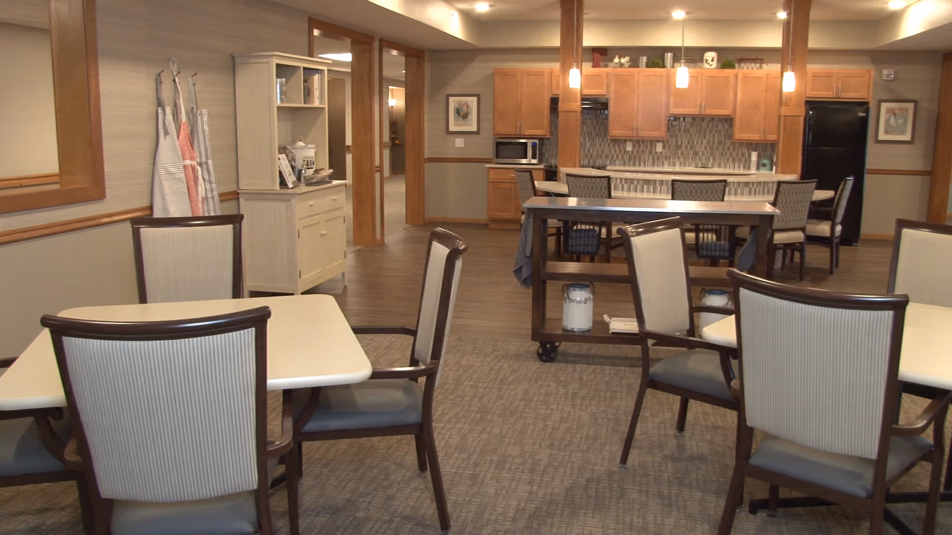 A spacious dining area in a senior living facility featuring multiple tables with chairs arranged around them. In the background, there is a kitchen area with wooden cabinets, a microwave, a refrigerator, and a kitchen island with chairs. Aprons hang on hooks on the left wall, and a small cabinet with shelves is also visible.