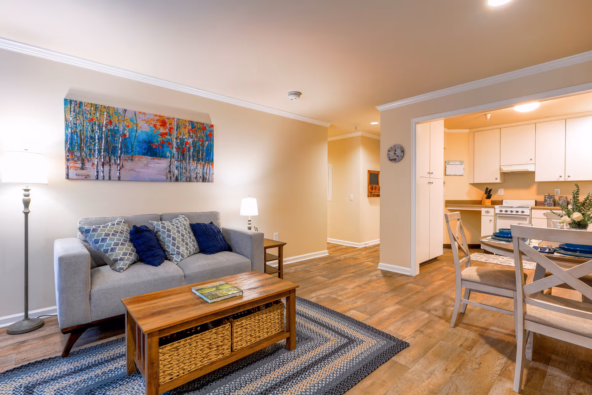 A cozy living room area with a gray sofa adorned with patterned and solid blue pillows, a wooden coffee table with woven baskets underneath, and a floor lamp. Above the sofa hangs a colorful painting of a forest with blue, red, and orange hues. To the right, there is a dining area with a wooden table set with blue plates and beige cushioned chairs. Beyond the dining area is a kitchen with white cabinets, a stove, and a countertop. The room has wood flooring and beige walls with white trim.