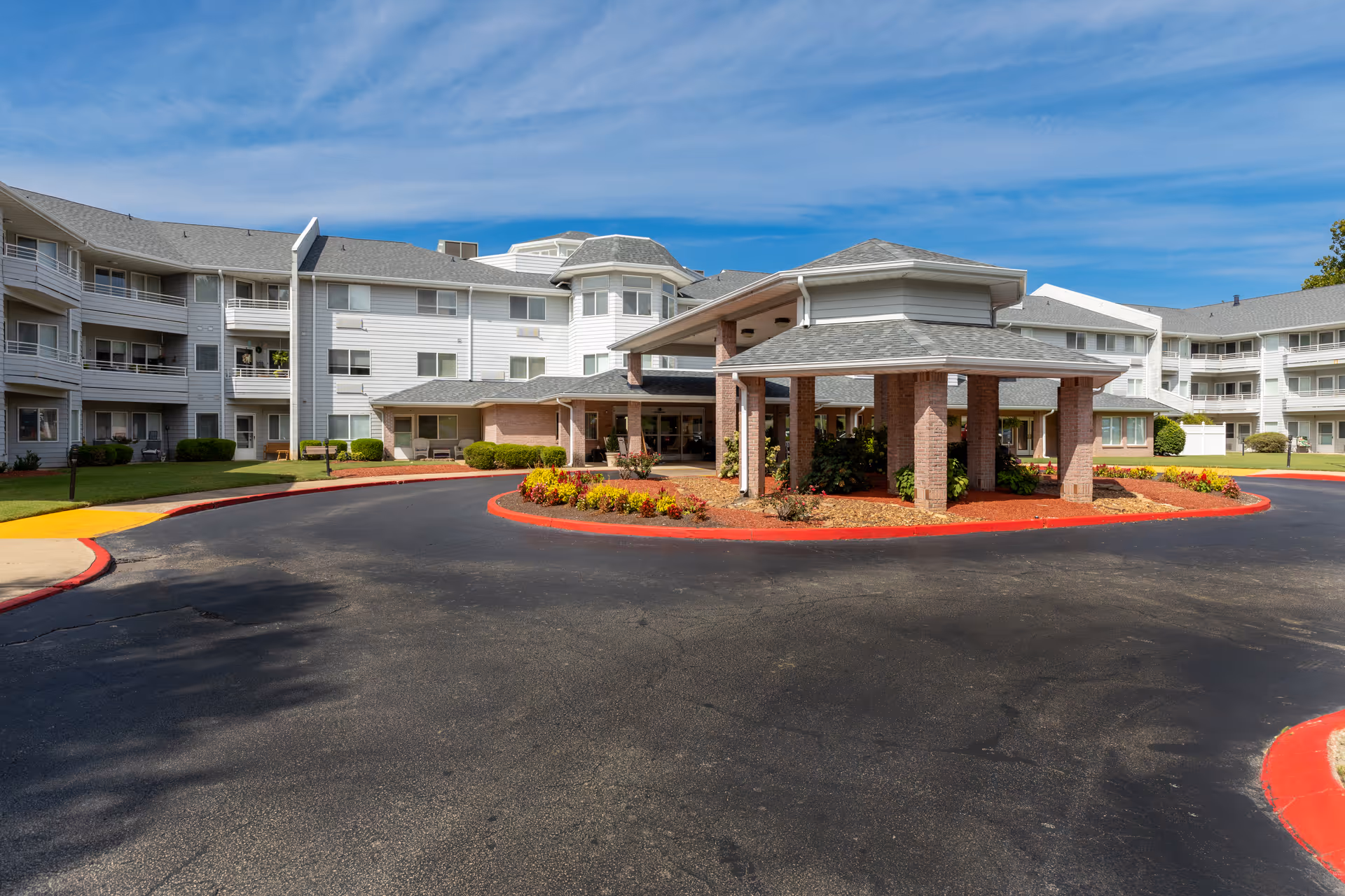 Exterior view of Jackson Meadow - A Provincial Senior Living Community, showing a large multi-story building with white siding and gray roofs under a blue sky. The entrance features a covered driveway with brick pillars and landscaped flower beds around it.