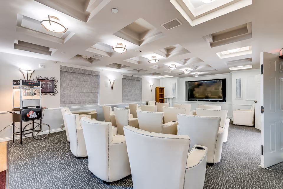 A bright screening room with rows of cream armchairs facing a large wall-mounted TV in a senior living facility.