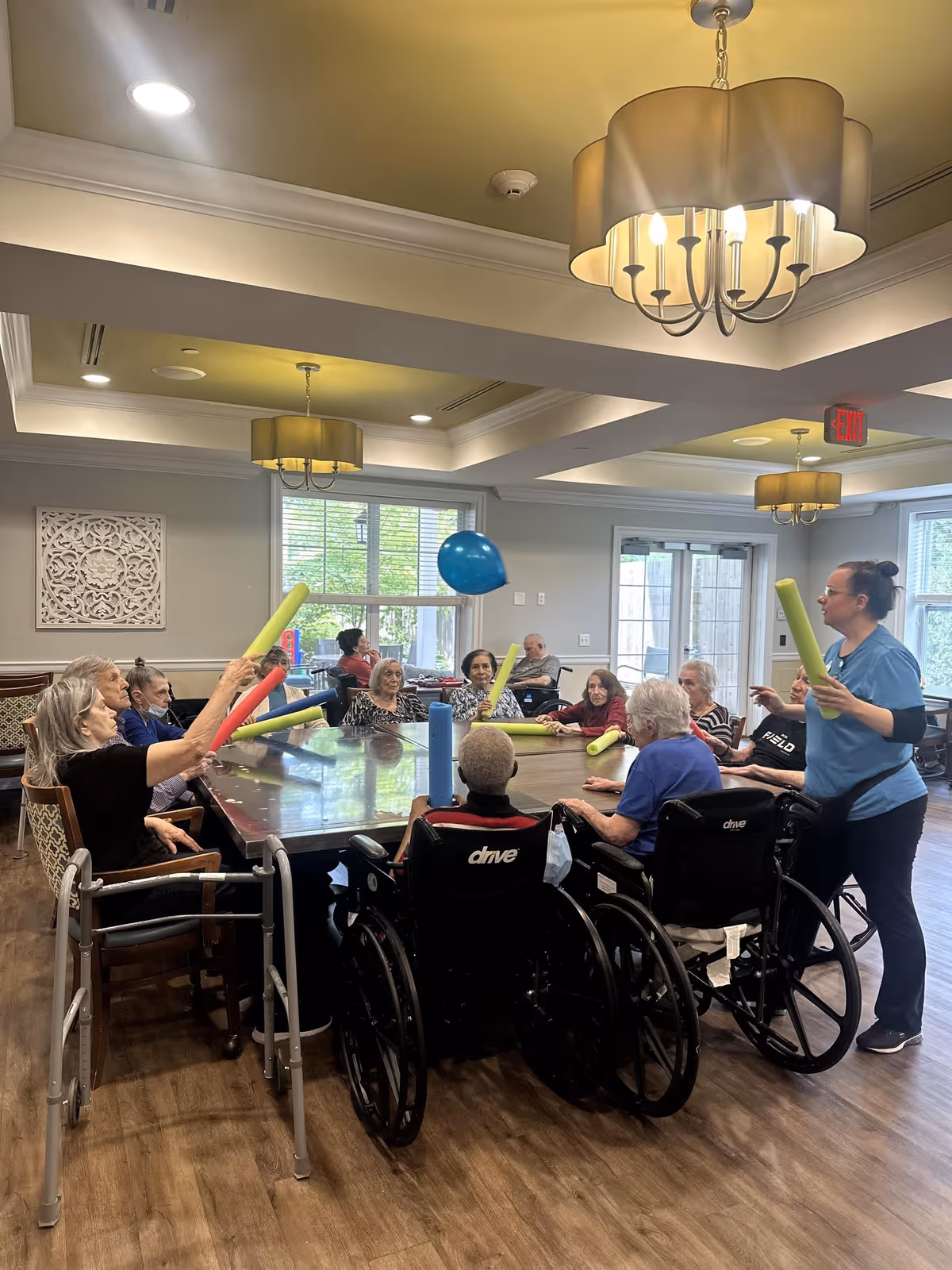 A group of elderly individuals, some in wheelchairs, seated around a large table in a well-lit room with modern ceiling lights. They are playing a game using colorful foam sticks to hit a blue balloon. A caregiver or staff member stands nearby, also holding a foam stick and engaging with the group. The room has large windows letting in natural light and light-colored walls with decorative wall art.