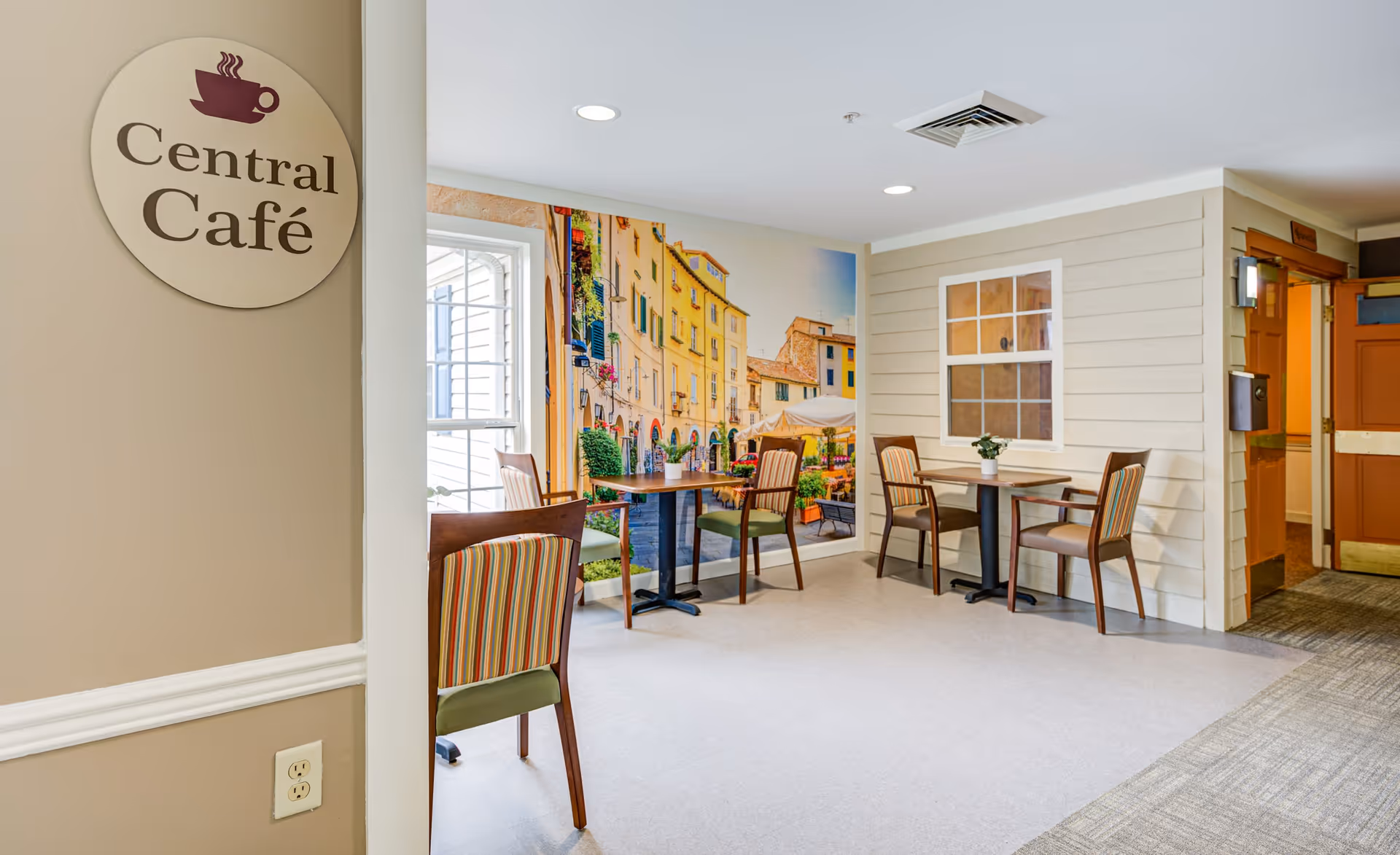 Interior view of a small café area named Central Café with two tables and four chairs. The walls are decorated with a large mural of a colorful European street scene and a window frame design. The floor is a combination of carpet and linoleum, and there is a doorway leading to another room.