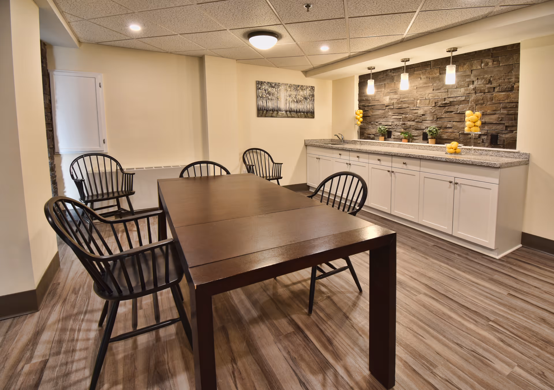 A dining room with a dark wooden table surrounded by black wooden chairs. The room features a countertop with white cabinets underneath and a stone accent wall behind it. Three pendant lights hang above the countertop, which is decorated with glass containers filled with lemons and small potted plants. The floor is wood-patterned, and a painting of trees hangs on the wall.