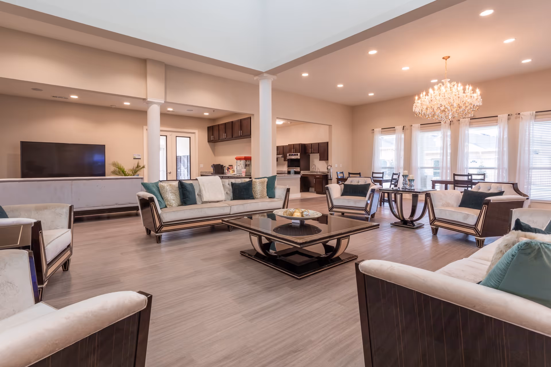A spacious and well-lit living room area with multiple beige and dark wood sofas and armchairs arranged around a central black coffee table. The room features light wood flooring, a large chandelier, recessed ceiling lights, and large windows with sheer white curtains. In the background, there is a kitchen area with dark wood cabinets and a dining area with tables and chairs.