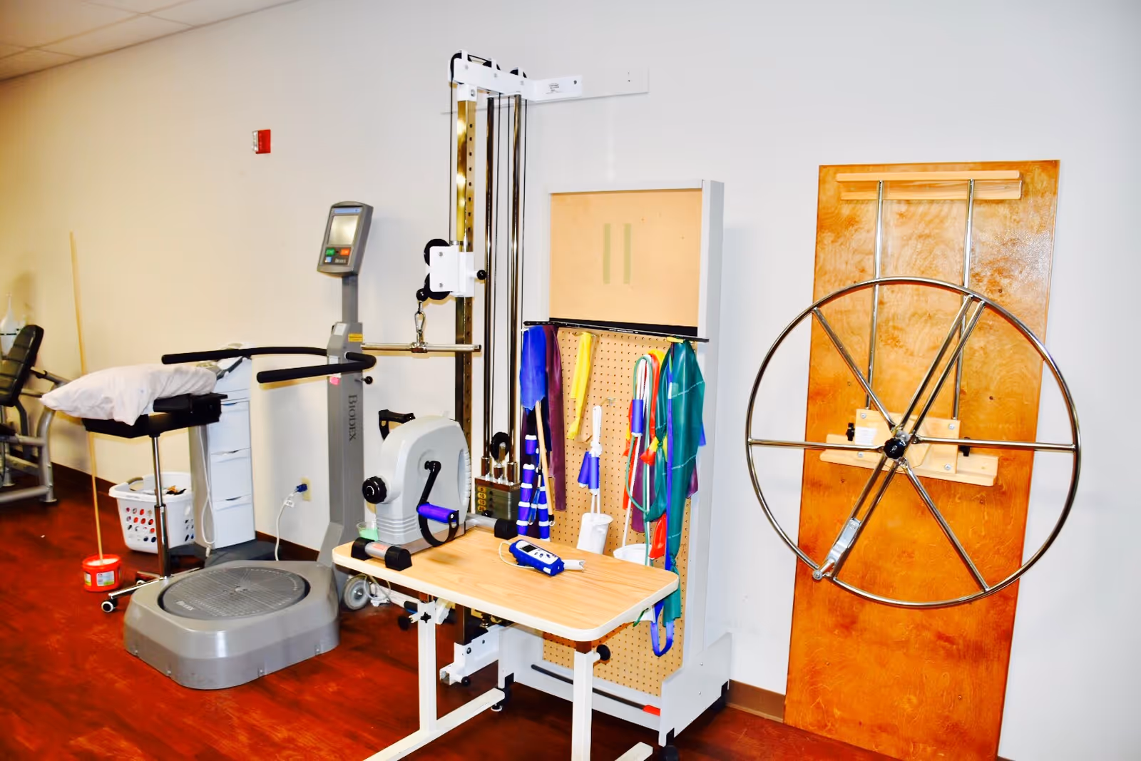 A physical therapy room with various rehabilitation equipment including a pulley system, resistance bands, a large metal wheel mounted on a wooden board, a vibration platform machine, and other exercise tools against a white wall with a wooden floor.
