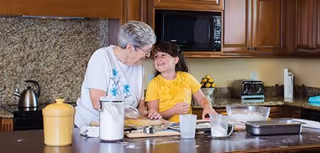 An elderly woman and a young girl smiling and interacting while preparing food together in a kitchen with wooden cabinets and granite countertops.