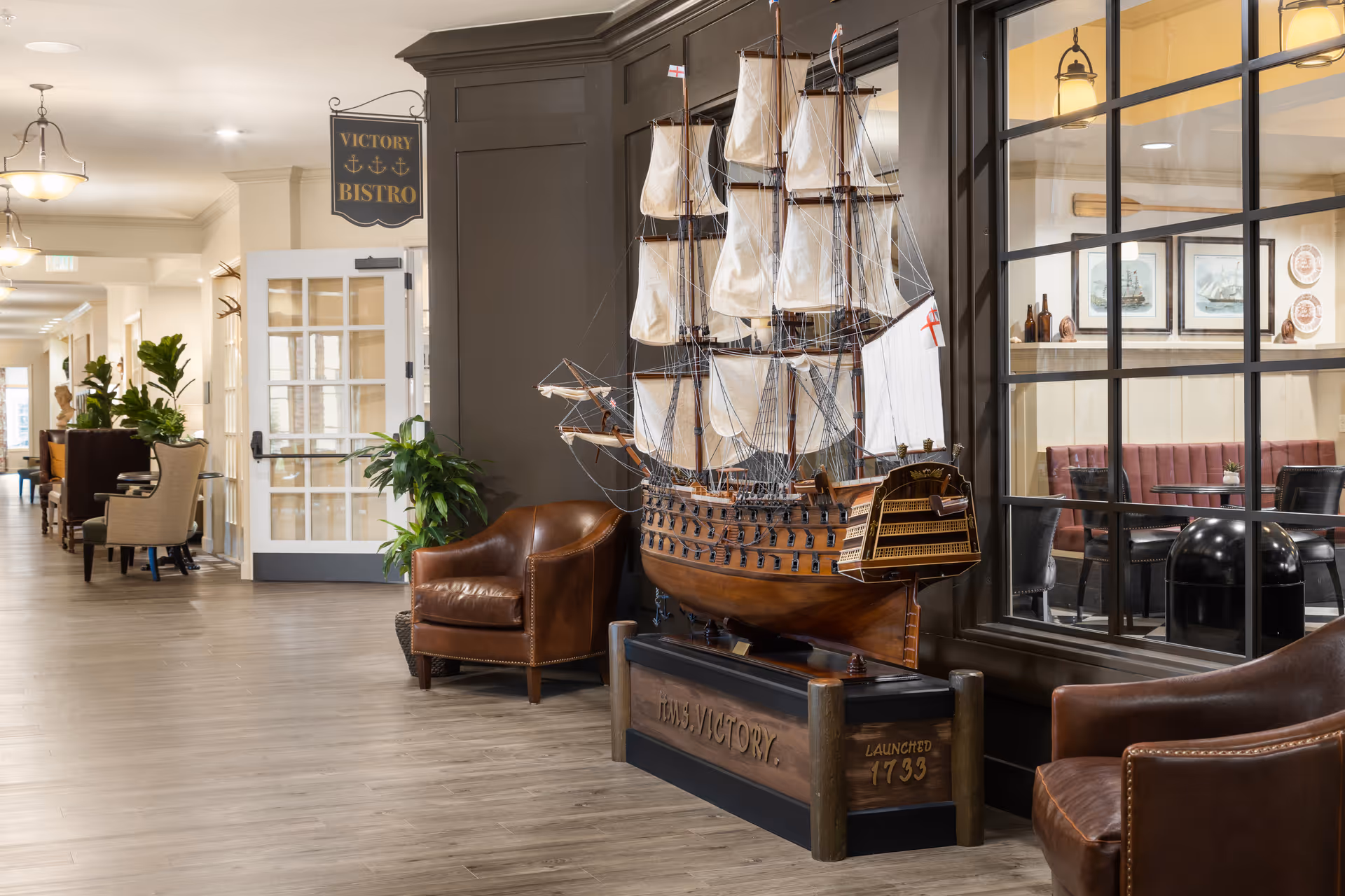 Interior hallway of The Park at Surprise Independent Living Community featuring a detailed model of the HMS Victory ship displayed on a stand between two brown leather chairs. A sign above reads 'Victory Bistro'. The hallway has light wood flooring, cream-colored walls, and several plants and seating areas along the corridor. Through a large window, a dining area with tables, chairs, and framed nautical artwork is visible.